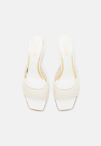 Zapatos slingback blancos con diseño de punta cuadrada, parte superior de tela y detalles dorados brillantes en la planta. Textura suave, acentos mínimos.