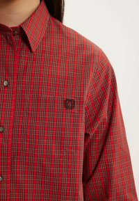 Chemise à carreaux rouges avec un col boutonné, dotée d'une poche et d'un petit logo brodé. Fabriquée en tissu léger avec une texture lisse.