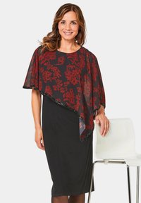 Zwart jurkje met een rode bloemenpatroon cape overlay. De cape heeft een doorzichtig textuur en een gegolfde rand. Model staat naast een witte stoel.