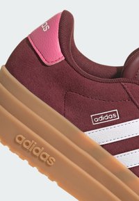 Maroon nubuk patika s bijelim prugama, ružičastim detaljima na peti i potplatom od gume. Sadrži Adidas logo sa strane.