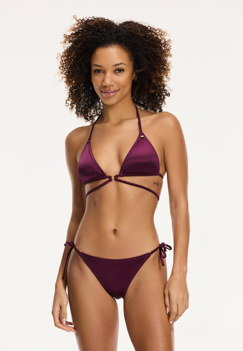 Bikini komplektas tamsiai vyšniniu atspalviu. Triangulinė liemenėlė su dirželių detalėmis ir auksinėmis akcentais; žemai įvyniotas apatinis trikotažas su raišteliais šonuose. Glotnus paviršius.
