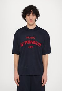 Jūras spēku acs sporta t-krekls ar sarkanu izšūtu tekstu "MILANO GYMNASIUM EA7". Brīvs piegriezums ar īsām piedurknēm un apaļu kakla izgriezumu.