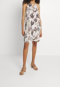 Kwiatowa sukienka na ramiączkach z dekoltem w kształcie łezki, z jasnoróżową podstawą i ciemnozielonymi liśćmi. Uzupełniona beżowymi sandałami na blokowym obcasie.