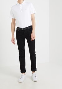 Polo bianco con colletto e patta a tre bottoni, abbinato a jeans neri e sneakers bianche. I jeans sono arrotolati alle caviglie.