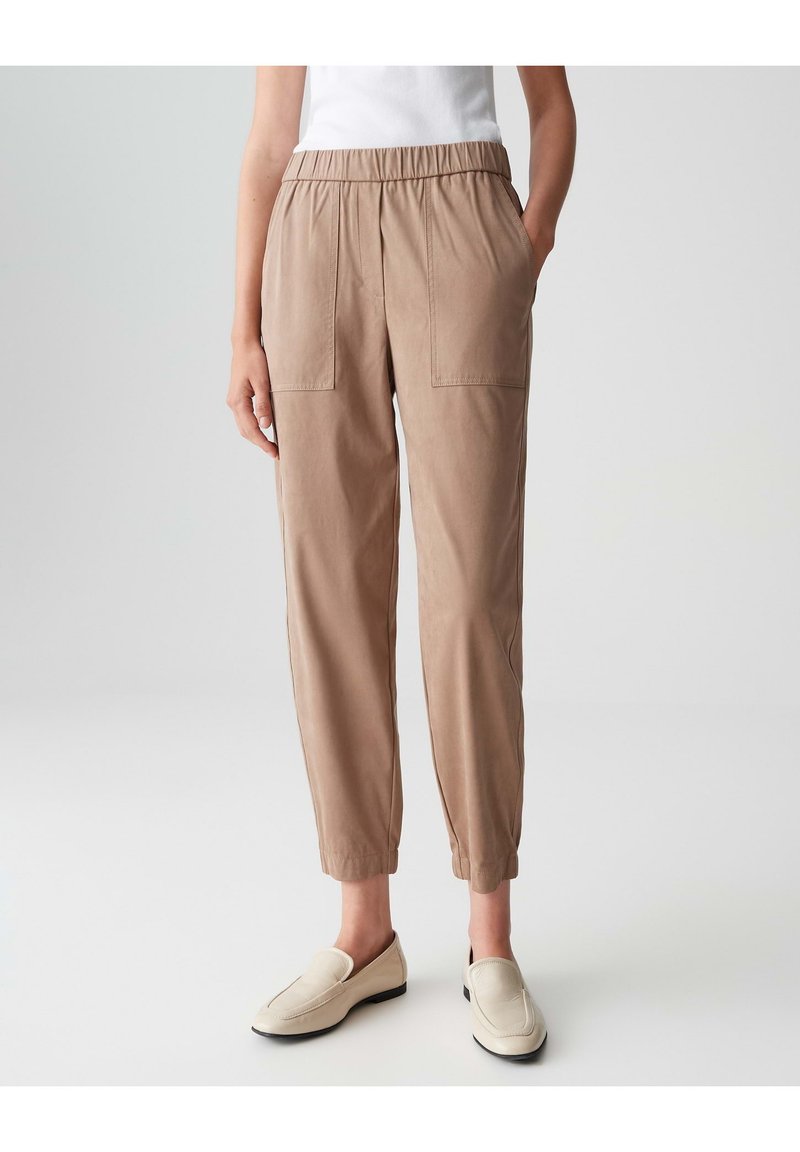 Hellbraune, elastische Hose mit zwei Vordertaschen, schmal zulaufenden Beinen und einer glatten Textur, kombiniert mit beigen Slippern.