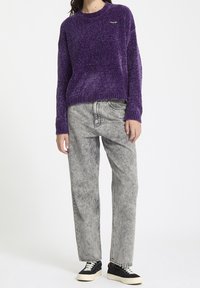 Personne portant un pull en tricot violet, un jean gris clair délavé à l'acide et des baskets noires à semelles blanches, debout devant un fond blanc.