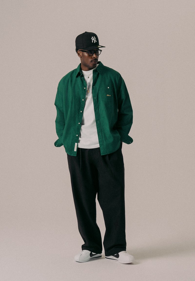 Camicia verde a bottoni sopra una maglietta bianca, abbinata a pantaloni neri larghi e sneakers bianche. Gli accessori includono un cappellino nero e occhiali.