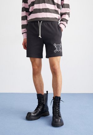 Pantalones cortos de sudor negro con cintura elástica y cordón, que cuentan con un bordado de calavera y huesos cruzados en la pierna izquierda. Botas de combate negras.