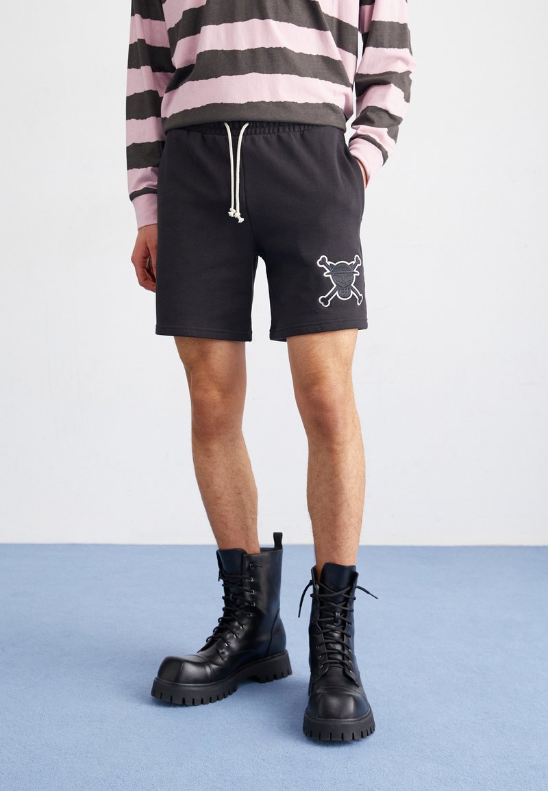 Pantalones cortos de sudor negro con cintura elástica y cordón, que cuentan con un bordado de calavera y huesos cruzados en la pierna izquierda. Botas de combate negras.
