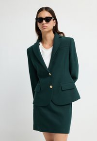 Mujer vestida con un blazer verde oscuro con botones dorados y una falda a juego, blusa blanca y gafas de sol cat-eye negras, de pie contra un fondo liso.