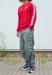 Persona che indossa un felpa rossa del Manchester United, pantaloni cargo grigi e scarpe nere e bianche, in piedi su una superficie pavimentata.