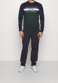 Marinblå och grön sweatshirt med vit text "SPORT" och Lacoste-logotyp, kombinerad med mörka byxor. Har rundad halsringning och ribbade ärmslut.