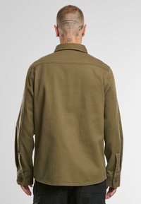 Haină-jachetă de culoare verde măslinie, cu mâneci lungi, guler cu butoni, material texturat și buzunare laterale. Cusături vizibile și accente cu nasturi.