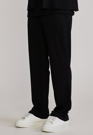 Alakehad mustades musta tekstuuriga pükstes ja valgete tennistega, seisavad ühtlase heleda halli tausta ees.