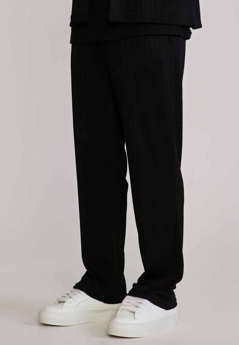 Alakehad mustades musta tekstuuriga pükstes ja valgete tennistega, seisavad ühtlase heleda halli tausta ees.