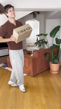 Caixa de cartão com o texto "zalando", segurada por uma pessoa vestindo uma camisa canelada castanha e calças de moletom cinzentas, de pé num chão de madeira.