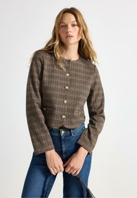 Brun ternet cropped jakke med lange ærmer og guldknapper, parret med blå højtaljede jeans. Struktureret stof med en veldefineret pasform.