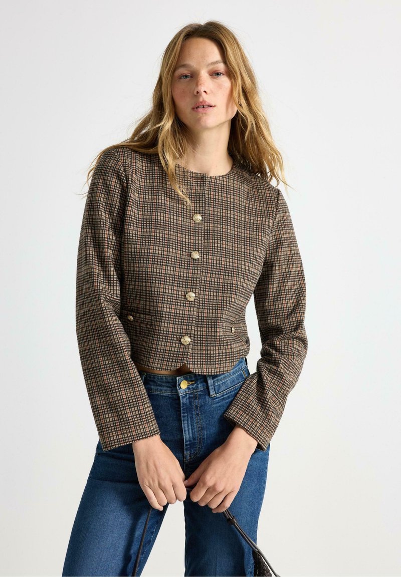 Brun ternet cropped jakke med lange ærmer og guldknapper, parret med blå højtaljede jeans. Struktureret stof med en veldefineret pasform.