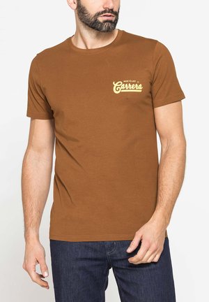 Uomo con barba che indossa una t-shirt marrone a maniche corte con il logo giallo "Made to Last Carrera" e jeans blu scuro, in piedi davanti a uno sfondo bianco.