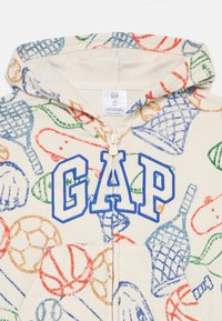 Giacca con cappuccio per bambini piccoli con motivi colorati di attrezzature sportive e grande logo GAP blu contornato sulla chiusura con cerniera frontale.