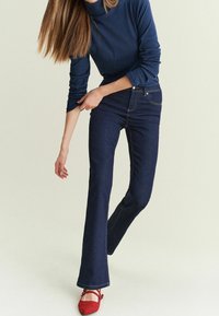Marineblå turtleneck-top parret med mørkeblå flared jeans, der har kontrasterende syninger og røde ballet-flade. Glat stof med en tætsiddende pasform.