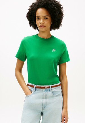 Kvinde med krøllet hår iført en grøn kortærmet skjorte og lyseblå jeans med et brunt bælte, stående med den ene hånd i lommen.