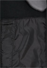Sort jakke med fleece-for, som har en glat sort nylon inderside, med en grå krave, synlige syninger og en lomme på venstre side.