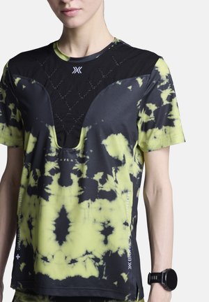 Persoon draagt een zwart-geel tie-dye sportshirt met korte mouwen, een mesh borstpaneel en een zwart sporthorloge om de pols.