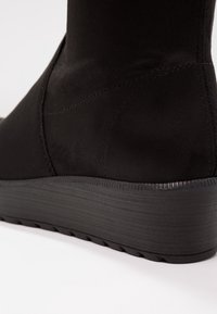 Svart ankelboot med en slät matt textur, utrustad med kilklack och subtila sömdetaljer längs sidan och hälen.
