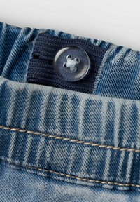 Cintura de mezclilla con fruncido elástico, con un botón transparente y tejido azul contrastante. Detalle de costura visible en los bordes.
