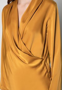 Musztardowożółta bluzka kopertowa z jedwabistego materiału z długimi rękawami. Posiada głęboki dekolt w kształcie litery V oraz drapowanie z przodu. Gładka, błyszcząca faktura.
