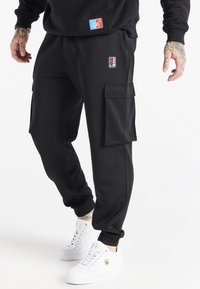 Mand iført sorte cargo-joggers med sidelommer og hvide høje sneakers, stående mod en hvid baggrund.