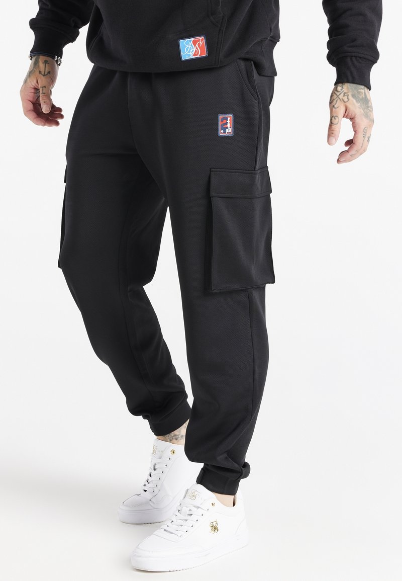Mand iført sorte cargo-joggers med sidelommer og hvide høje sneakers, stående mod en hvid baggrund.