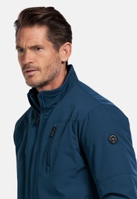 Marineblauwe softshell jas met een hoge kraag, een ritszak op de borst en een drukknooplogo op de linkermouw. Glad textuur, aansluitend ontwerp.