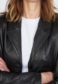 Schwarze Lederblazer mit Reverskragen, Ein-Knopf-Verschluss und subtilen Nähdetails, über einem weißen Rundhalsausschnitt-T-Shirt getragen.