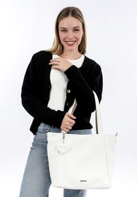 Bolso tote de cuero blanco con un acabado texturizado, asas cortas en crema y un charm en forma de corazón. La modelo lleva un cárdigan negro y unos vaqueros.