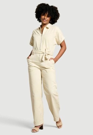 Frau mit lockigem Haar, die einen cremefarbenen Jumpsuit mit Gürtel und beige Sandalen mit Absatz trägt, steht mit den Händen in den Taschen.