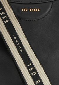 Sac en cuir noir avec une surface texturée, accompagné d'une sangle crème contrastante portant l'inscription "TED BAKER LONDON" en noir et un détail de logo doré.