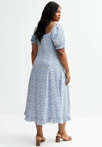 Robe fleurie bleu clair avec de courtes manches bouffantes, un ourlet à volants et une ouverture au dos. Associée à des sandales à talons marron.