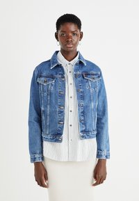 Veste en denim bleu moyen avec un col classique, des poches avant et des boutons. Portée sur une chemise rayée blanche et grise.
