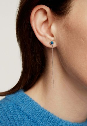 Primer plano de la oreja de una mujer que lleva un pendiente plateado en forma de estrella con una gema azul y una delicada cadena larga que cuelga.