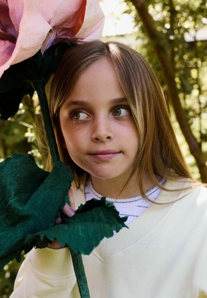 Jeune fille aux longs cheveux bruns tenant une grande fleur en papier rose, regardant sur le côté, en plein air avec des arbres verts sous une lumière douce.
