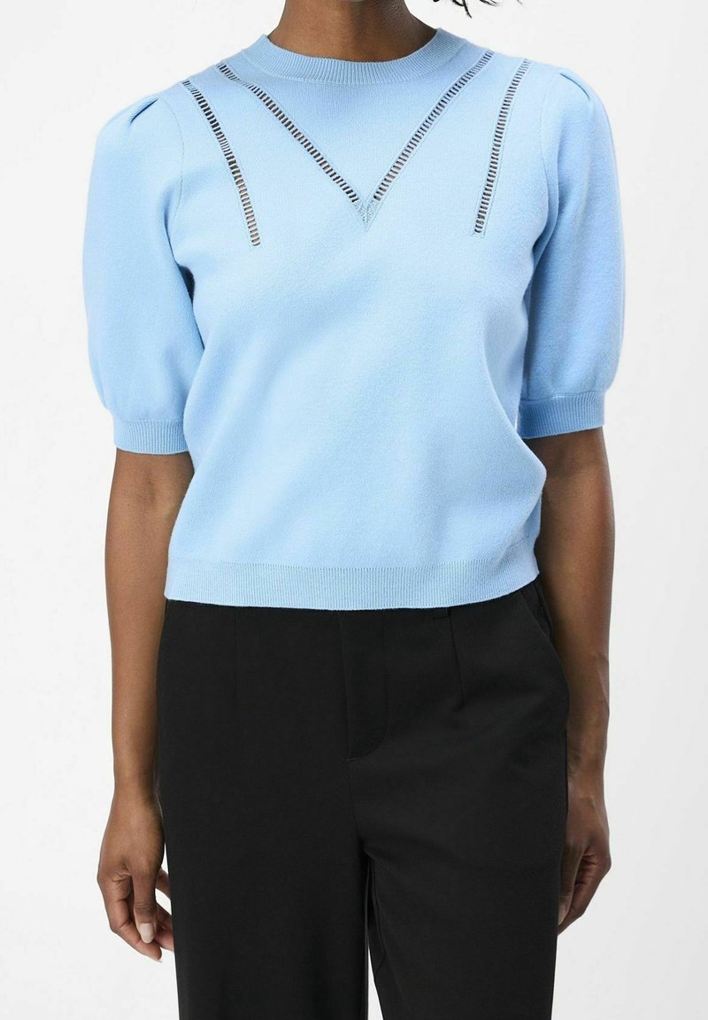 Femme portant un pull bleu clair à manches courtes avec des détails découpés près de l'encolure et un pantalon noir taille haute.