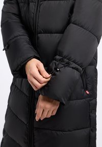 Czarna kurtka puchowa z poziomym pikowaniem, z przednim zamkiem błyskawicznym i regulowanymi mankietami na troczki. Czerwona metka widoczna z boku.