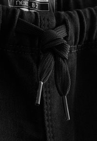 Gros plan sur un pantalon en tissu noir avec un cordon noir noué doté d'embouts métalliques et une partie d'une étiquette cousue au-dessus de la ceinture.