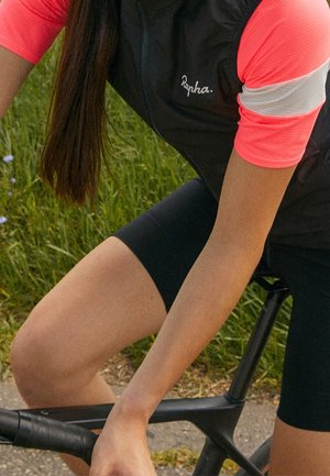 Černá cyklistická vesta s růžovými a bílými krátkými rukávy, přiléhavý střih a měkká látka. Jezdec drží černá řídítka kola na pozadí zelené trávy.