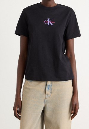 Person iført en sort Calvin Klein t-shirt med lilla og pink logo, kombineret med lyse beige denimjeans.