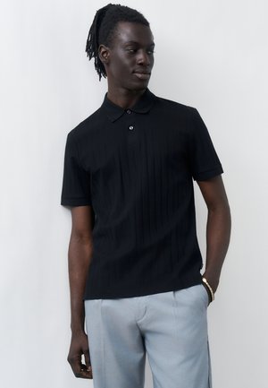 Giovane uomo con dreadlocks che indossa una polo nera a maniche corte e pantaloni grigio chiaro, in piedi su uno sfondo chiaro e uniforme.