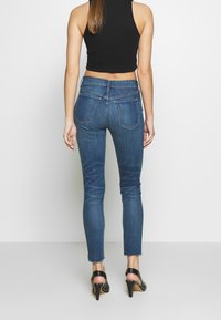 Jeans ajustados en denim azul oscuro con bolsillos traseros, dobladillo deshilachado y un diseño entallado, combinados con sandalias de tacón negras.