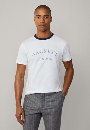 Camiseta de algodón blanca con cuello de punto en navy, que presenta el texto "HACKETT LONDON" en azul. El modelo la lleva con pantalones gris de rayas.